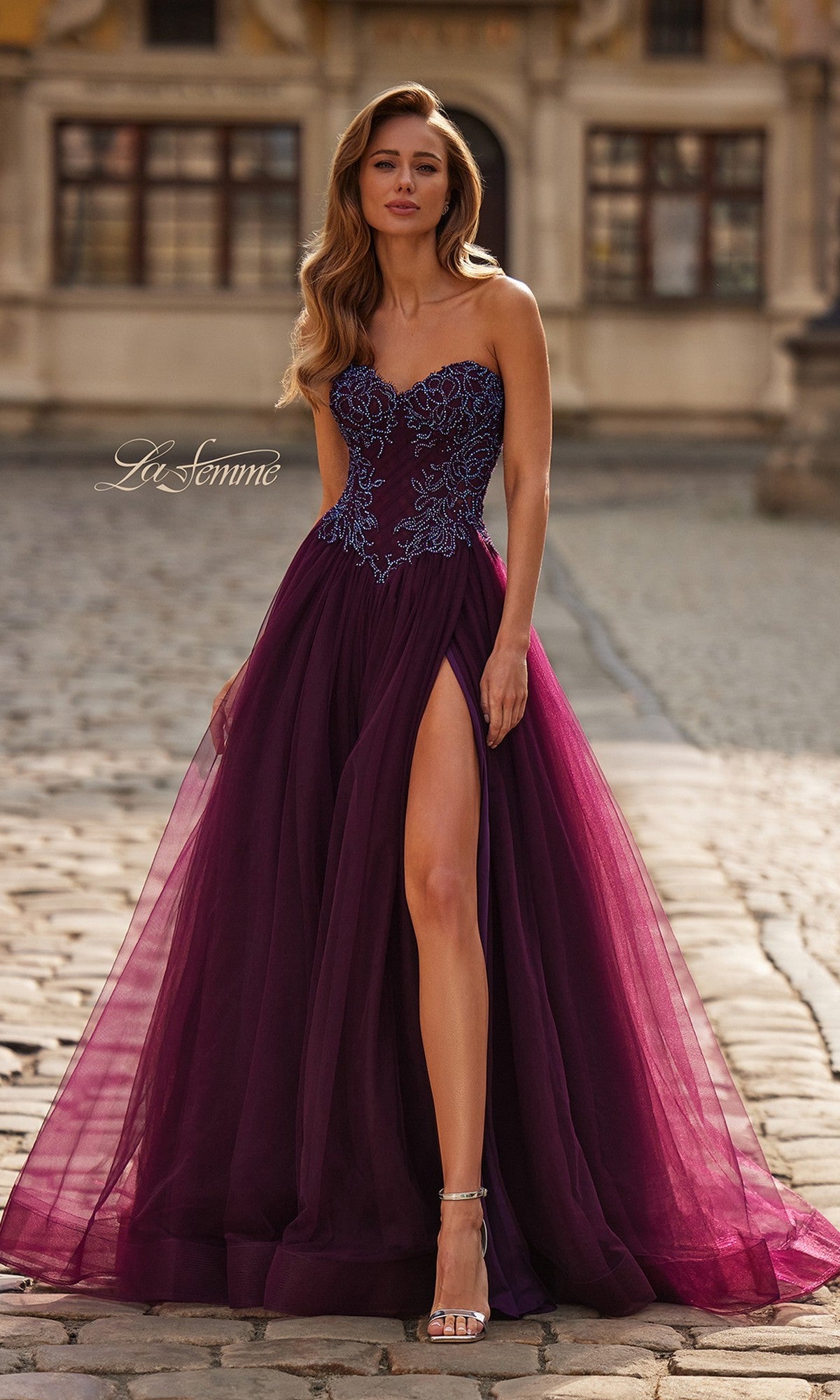 A woman in the La Femme 33410 long prom dress in dark berry, featuring a strapless beaded corset bodice, floral-embroidered top, tulle A-line skirt, and high leg slit, stands on a cobblestone path.